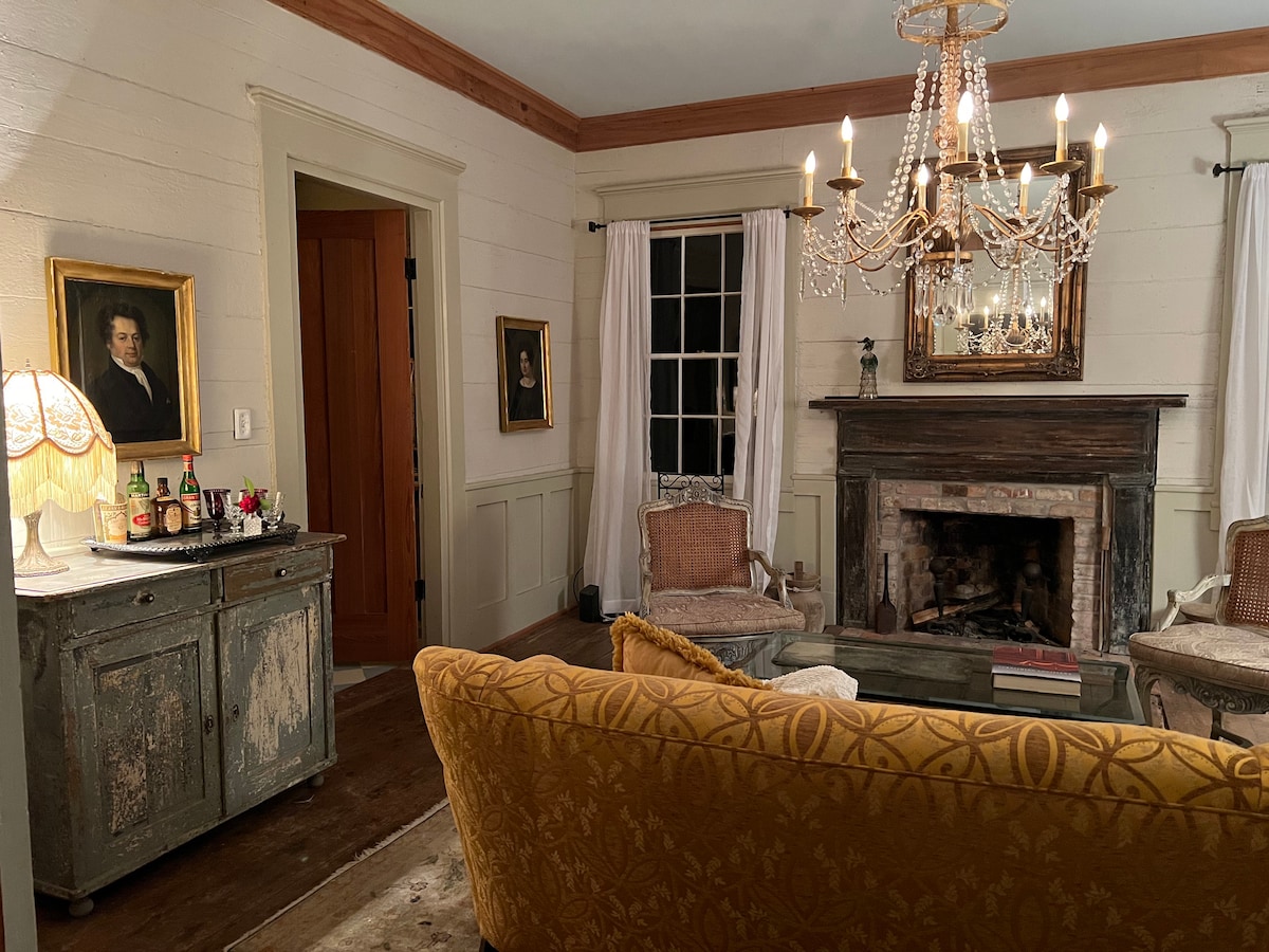 The living area features an elegant design, highlighted by a classic chandelier above a comfortable sitting arrangement. A vintage fireplace, framed artworks on the walls, and a lamp beside a console table contribute to the room's historic charm.