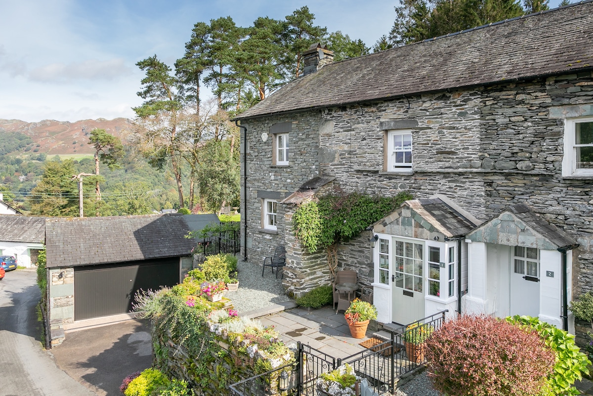 The traditional stone cottage is framed by greenery, showcasing charming architectural details. A small patio area with potted plants sits at the entrance. Surrounding hills provide a scenic backdrop, enhancing the cottage's tranquil setting on a quiet lane.