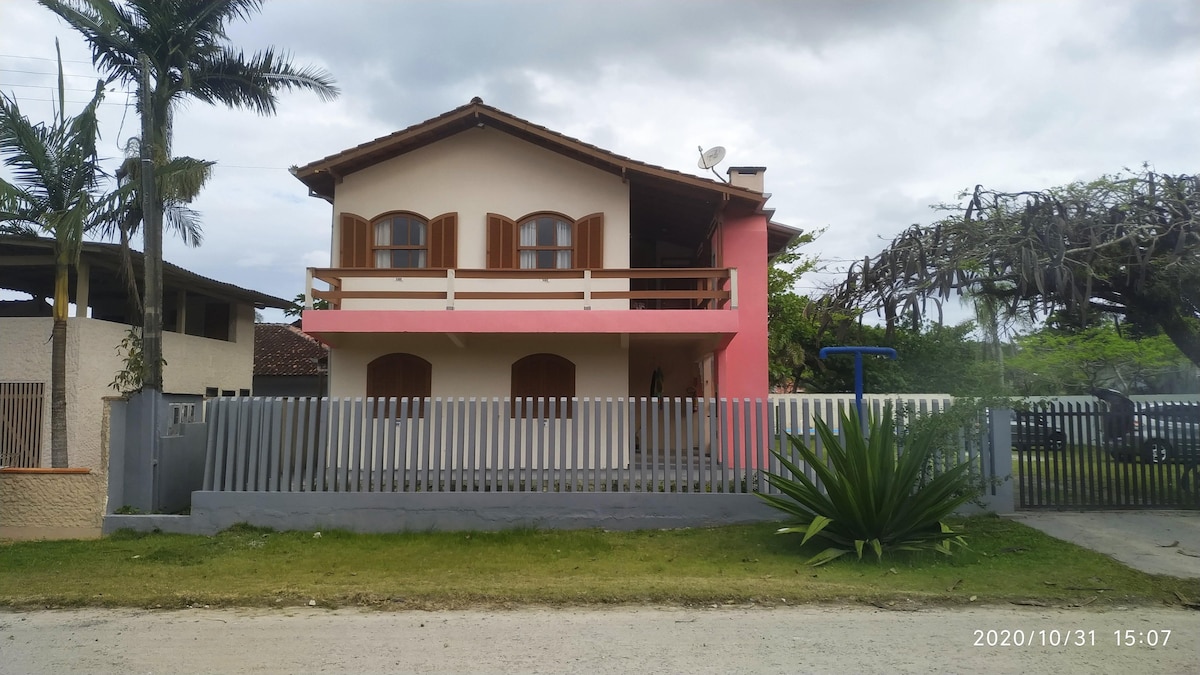 A two-story building features a mix of light pink and beige exterior, with wooden shutters on the windows. A balcony is visible on the upper floor, and a simple wooden fence encloses the front yard, which is landscaped with grass and tropical plants.