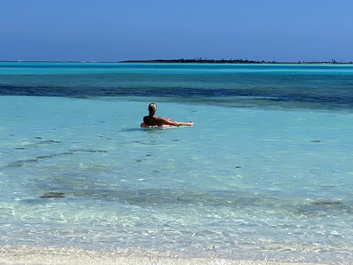 A tranquil seascape features a calm turquoise sea reflecting bright blue skies. A figure is seen seated in the water, surrounded by gentle waves. The sandy beach softly transitions into the clear water, creating a serene atmosphere along the shore.