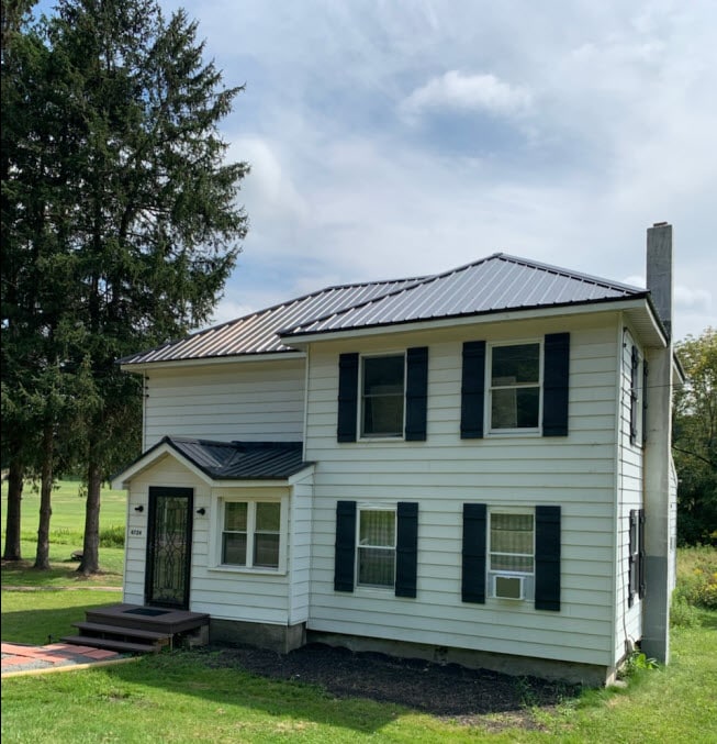 A charming farmhouse is showcased with a white exterior, complemented by dark shutters. The metal roof adds a modern touch. Green grass surrounds the house, which is situated near tall trees and offers a view of the golf course.