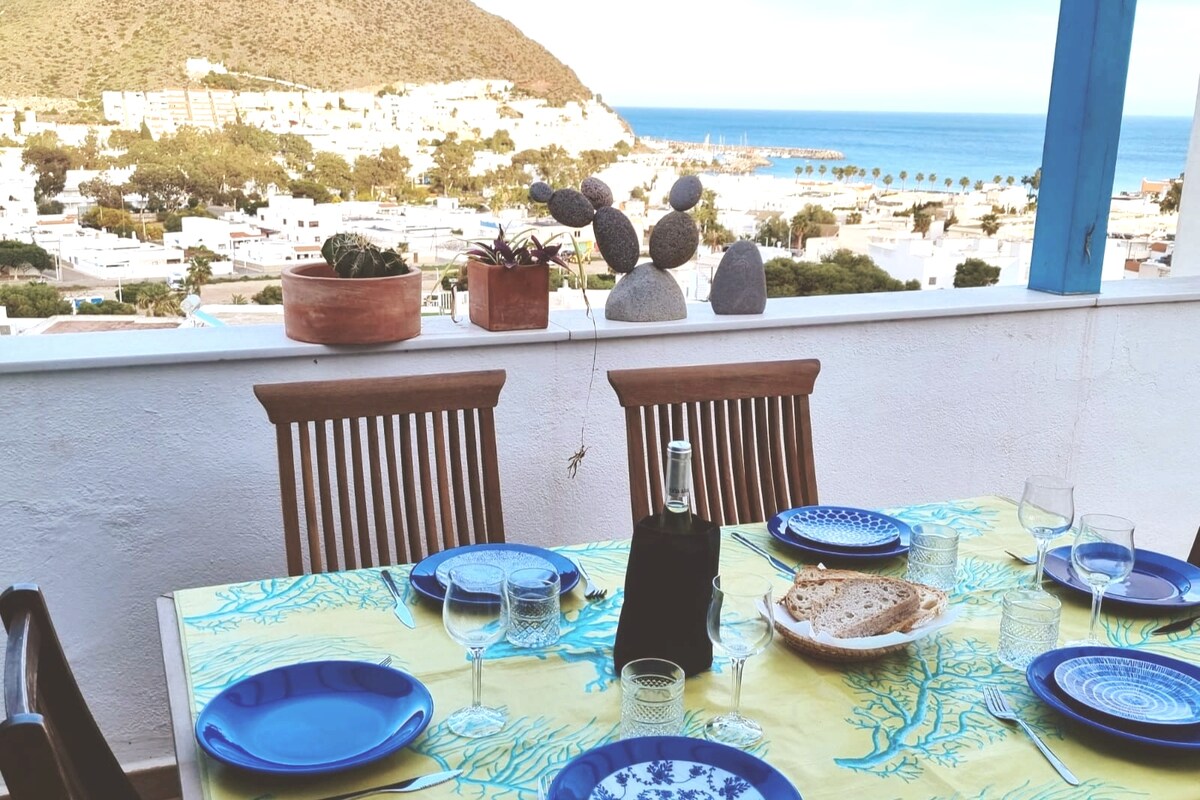 A dining table is set on a balcony, featuring blue plates and clear glasses. Natural light highlights a coastal view with mountains in the background. Potted plants and stacked stones add to the relaxed atmosphere, creating an inviting space for meals.