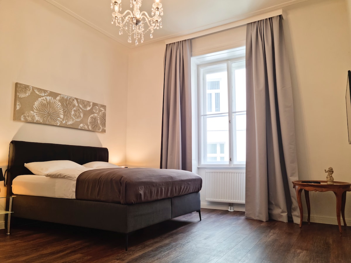 A well-lit bedroom features a comfortable bed with neutral bedding and decorative wall art. A window draped with light curtains allows natural light to illuminate the space. An elegant chandelier hangs from the ceiling, adding a touch of sophistication.