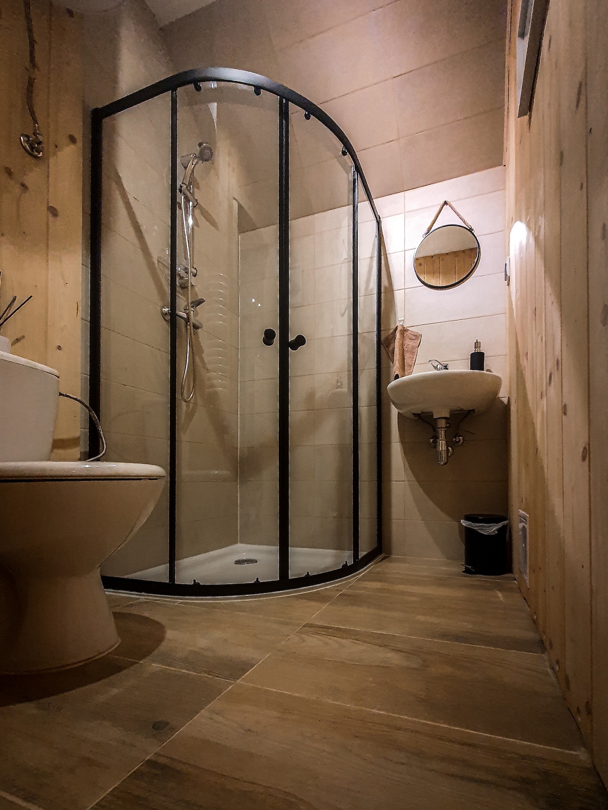The bathroom features a modern shower with a glass enclosure, a toilet, and a small sink. Light wood paneling and neutral tiles create a calming environment. A round mirror hangs on the wall, enhancing the practicality of the space.
