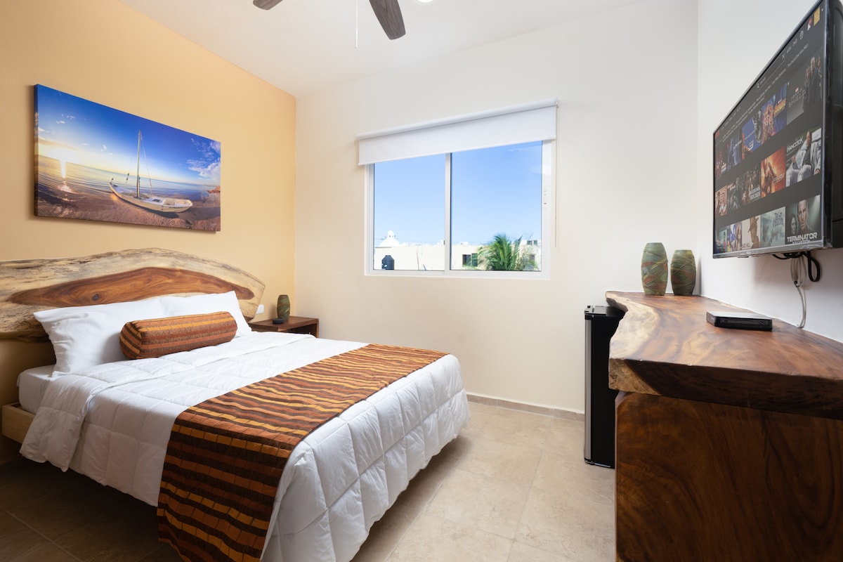 A well-lit bedroom features a spacious bed with a textured blanket across the bottom. A wooden headboard and side table complement the room's decor. A large window allows natural light to fill the space, and a wall-mounted Smart TV offers entertainment options.