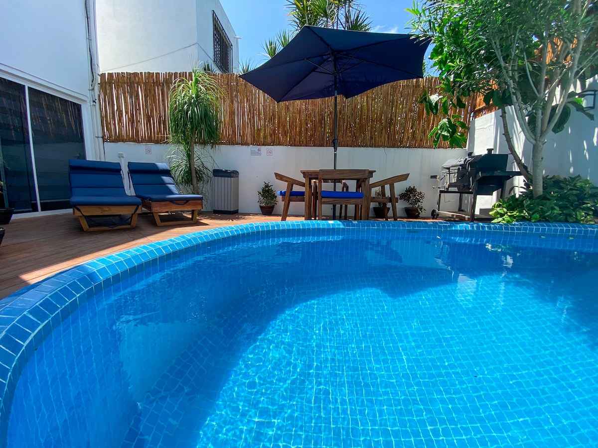 A refreshing outdoor pool is surrounded by a wooden deck and lush greenery. Two lounge chairs are positioned under a large umbrella, offering shade. A dining table and grill area can be seen, creating a welcoming space for relaxation and outdoor dining.