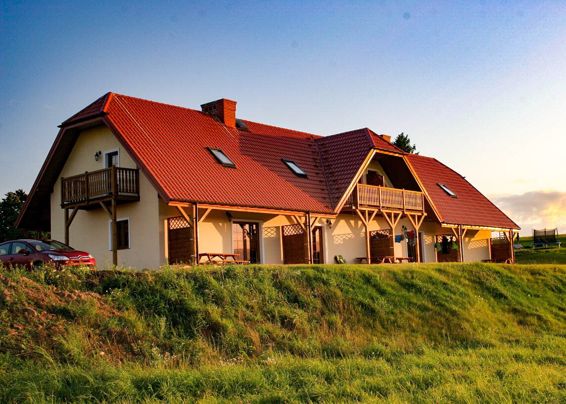 The exterior of a spacious building is shown, featuring a red-tiled roof and balconies. Lush green grass surrounds the structure, with a few picnic tables visible on the patio. A car is parked nearby, and the landscape stretches out into the distance.