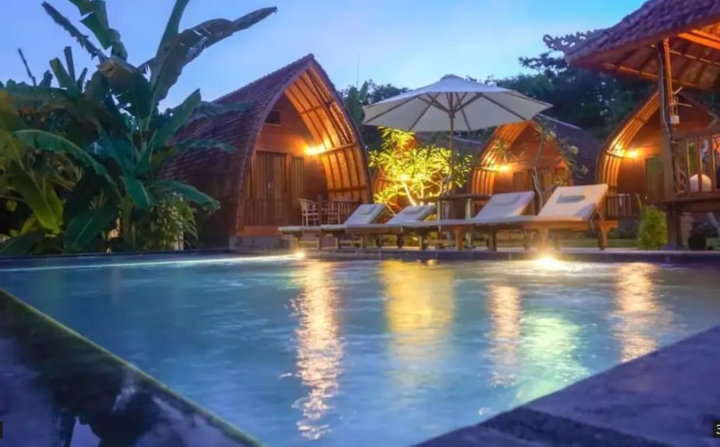 A serene swimming pool is framed by lounge chairs, surrounded by lush greenery and unique hut-like structures in the background. The pool water reflects the ambient lighting, creating a calming atmosphere as twilight descends.