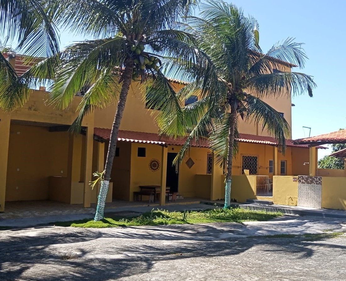 The exterior of the yellow duplex is framed by tall palm trees, creating a tropical ambiance. A covered patio area is visible, with outdoor seating partially obscured in the shade. The entrance shows a welcoming view into the home, highlighting its spacious design.