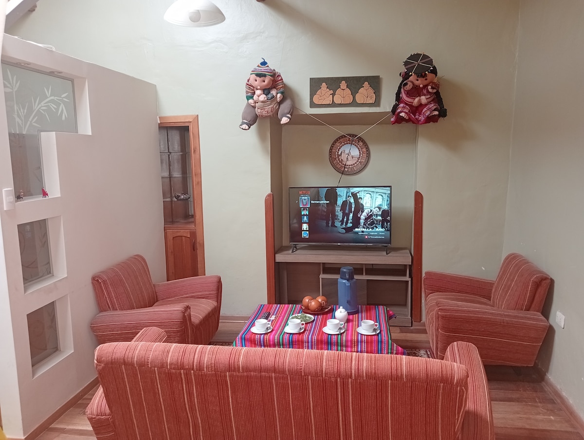 A cozy living area features a television mounted on the wall, accompanied by a wooden media console. Two striped armchairs and a sofa, adorned with Andean decor, create a welcoming seating space. A colorful table is set with traditional cups and a teapot, enhancing the room's cultural charm.