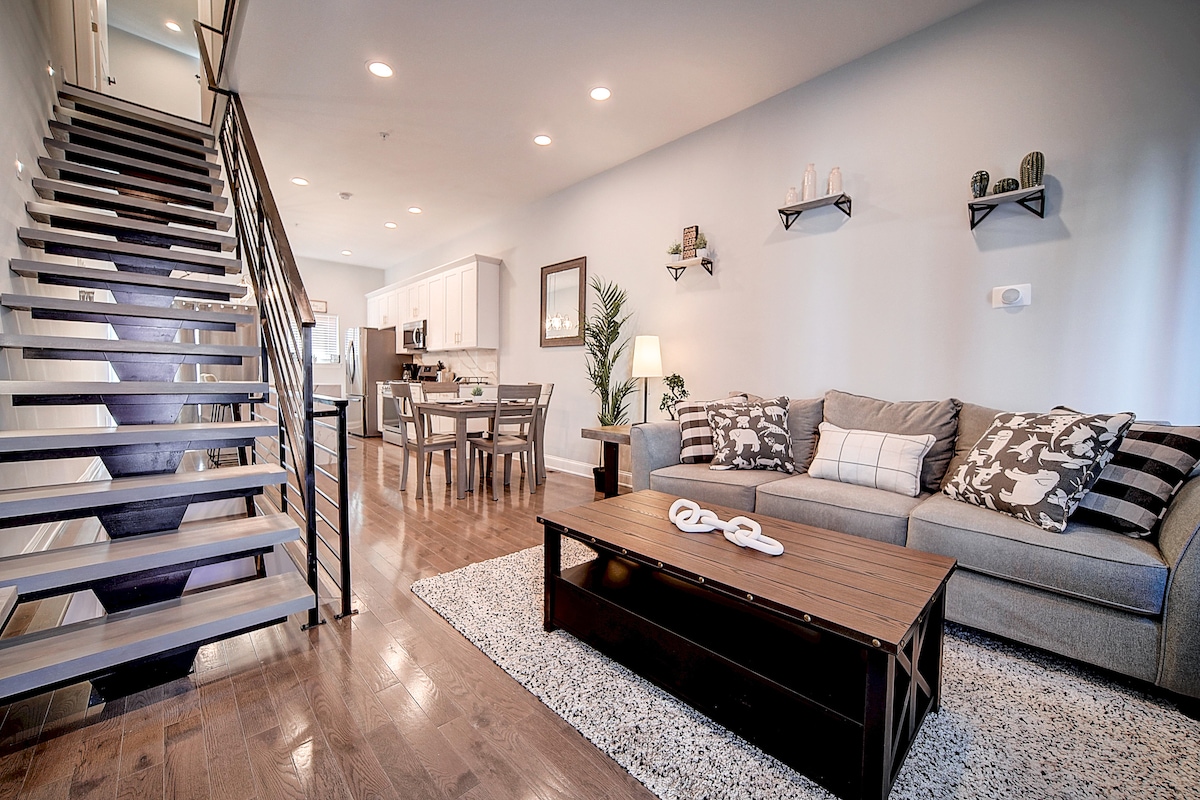 A spacious living area features a gray sofa adorned with decorative pillows, set on a cozy rug. A wooden coffee table sits in front, and a dining area is visible in the background. Light from recessed ceiling fixtures brightens the open layout, which connects to a staircase.