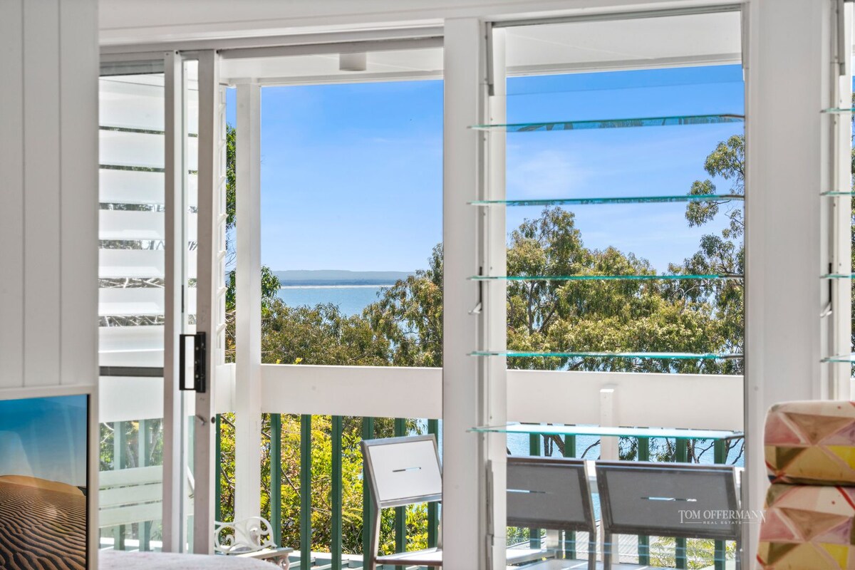 Large glass doors create a connection to the outside, allowing abundant natural light within. The view overlooks lush greenery and the bay beyond, framed by elegant louvered windows that offer ventilation while maintaining a sense of openness.