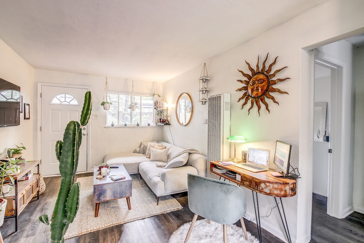 A light-filled living area showcases a comfortable sectional sofa paired with a rustic coffee table. Sunlight streams through the window, highlighting wall decor and a stylish desk with a chair. A tall cactus adds a touch of greenery to the space, enhancing the overall atmosphere.