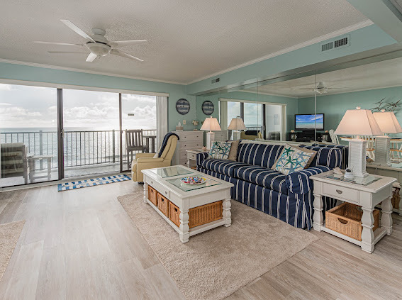 The spacious living area features a striped sofa and a glass-top coffee table, surrounded by light-colored furnishings. Large windows provide unobstructed ocean views, while soft lighting from table lamps creates a cozy ambiance. A comfortable chair and an area rug complete the inviting space.