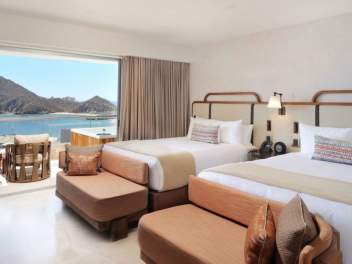 A spacious room is displayed, featuring two queen beds with wooden frames. A large private balcony is visible through a collapsing glass wall, showing views of the Sea of Cortez. A comfortable seating area is positioned near the entrance, enhancing the room's inviting ambiance.