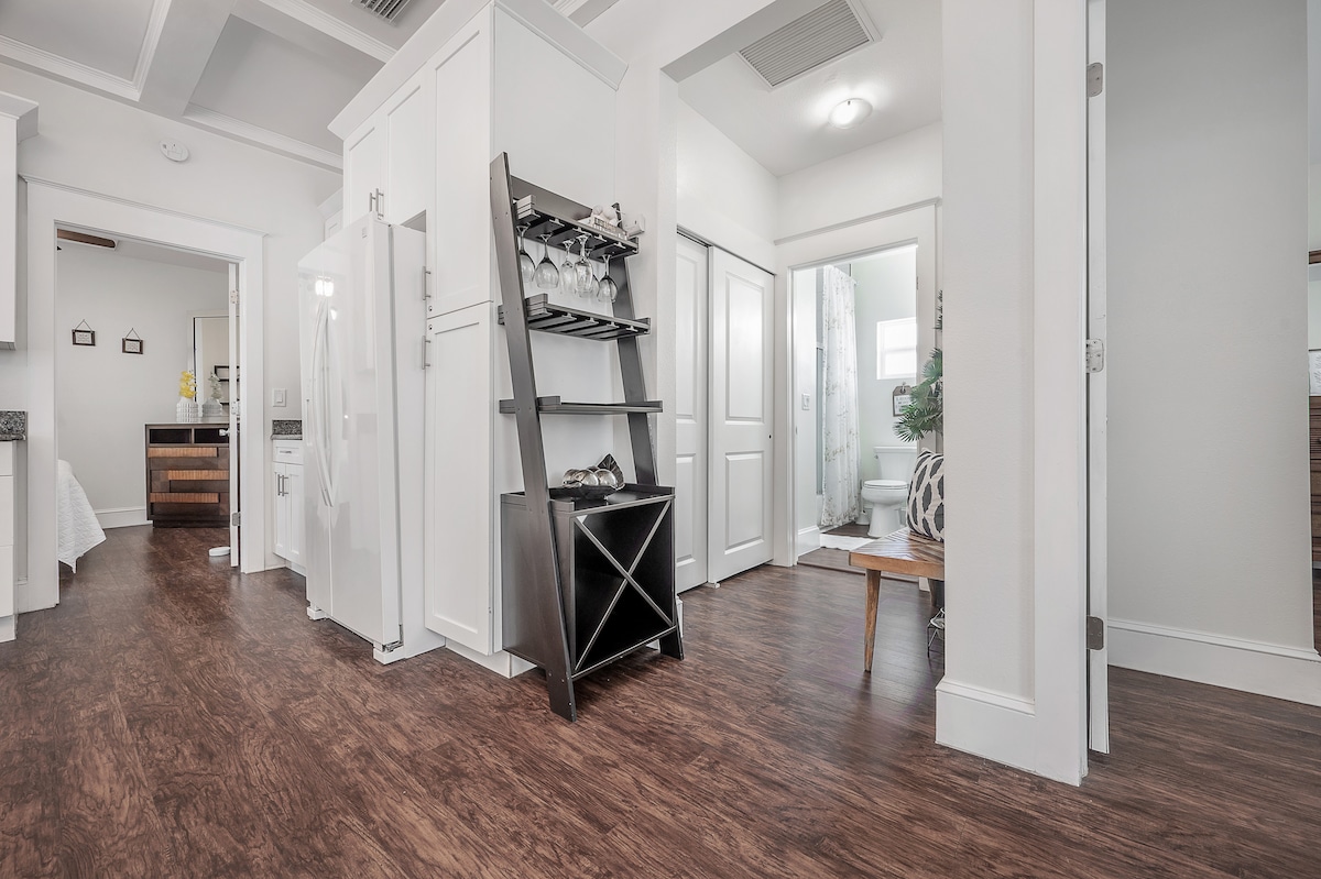 A spacious entryway features wooden flooring, leading to various rooms. A sleek, black shelving unit holds glassware, while a white refrigerator is visible on the left. Doorways mark entrances to adjacent rooms, including an open bathroom partially seen in the distance.