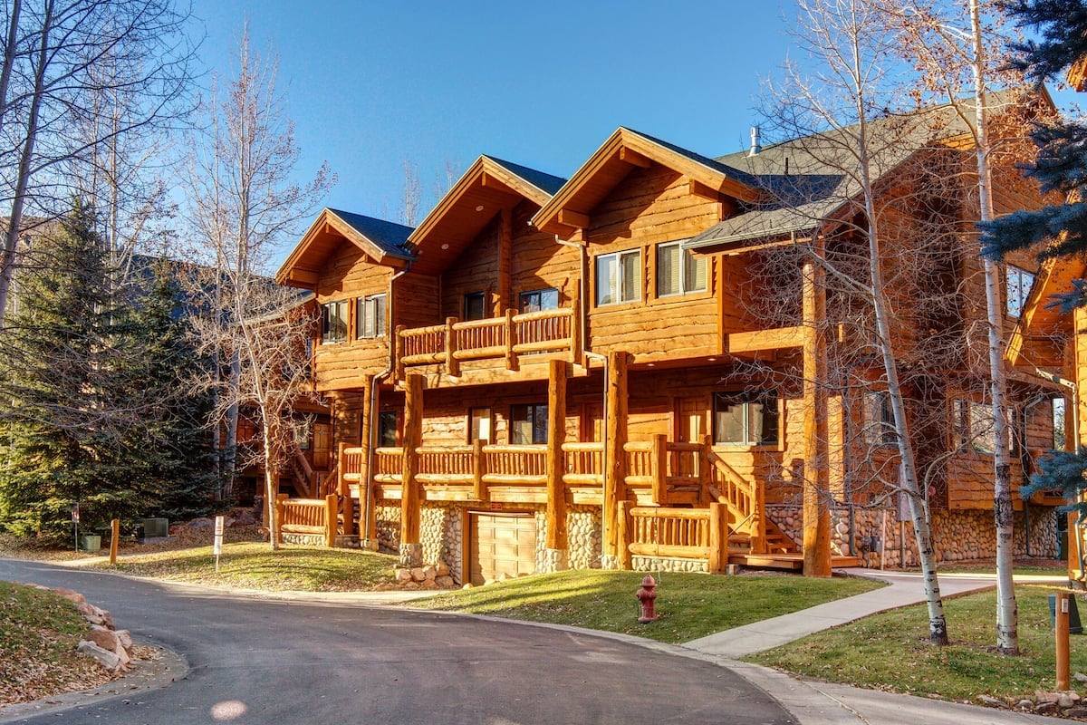 A rustic wooden condo is shown, featuring multiple balconies with log railings. The structure is set among tall trees, providing a natural setting. A paved path leads to the entrance, with a driveway and green lawn visible in front.
