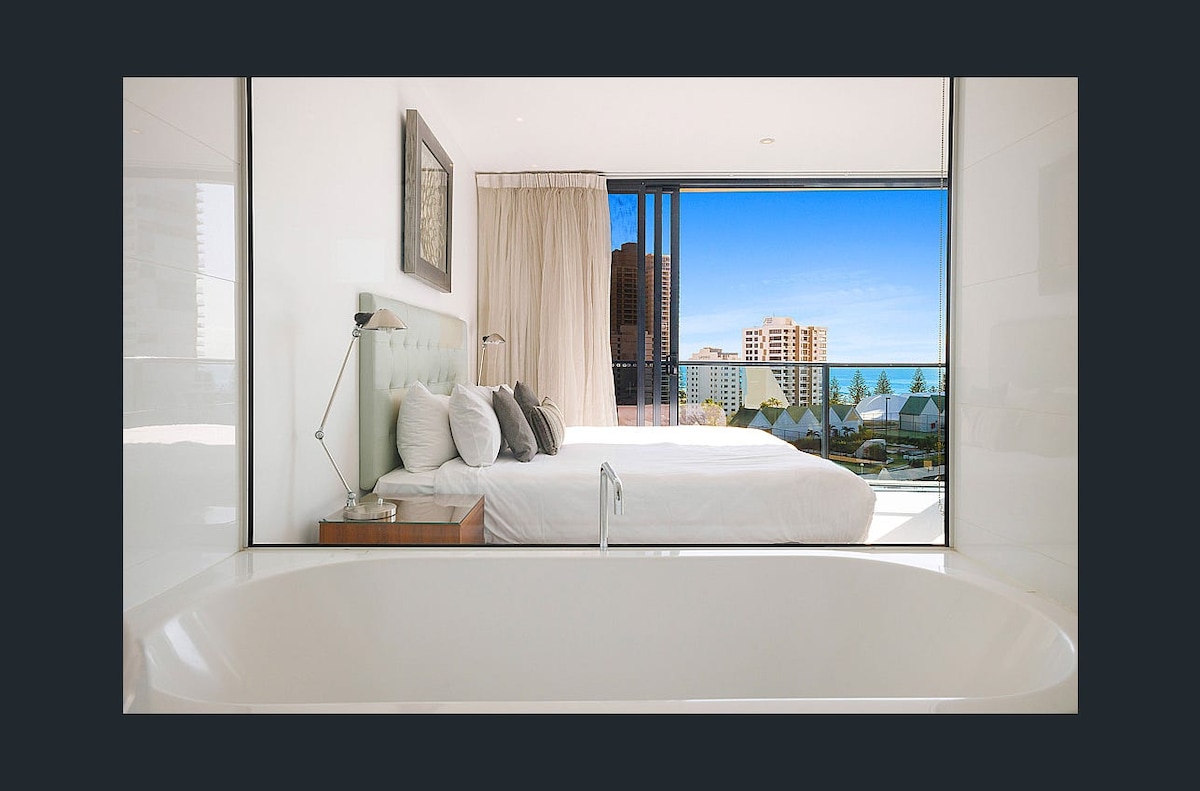 A modern bedroom is visible through a large glass window, showcasing a spacious bed adorned with soft pillows. Natural light fills the room, enhancing the view of the skyline and ocean beyond. A sleek bathtub is positioned in the foreground, offering a glimpse of relaxation.