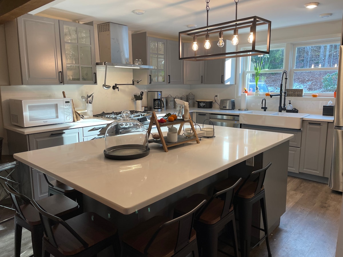 A spacious kitchen is equipped with modern appliances, including a microwave and coffee maker. The large island, featuring a white quartz countertop, offers seating for eight. Pendant lights hang above, and windows provide natural light, enhancing the functional and inviting space.