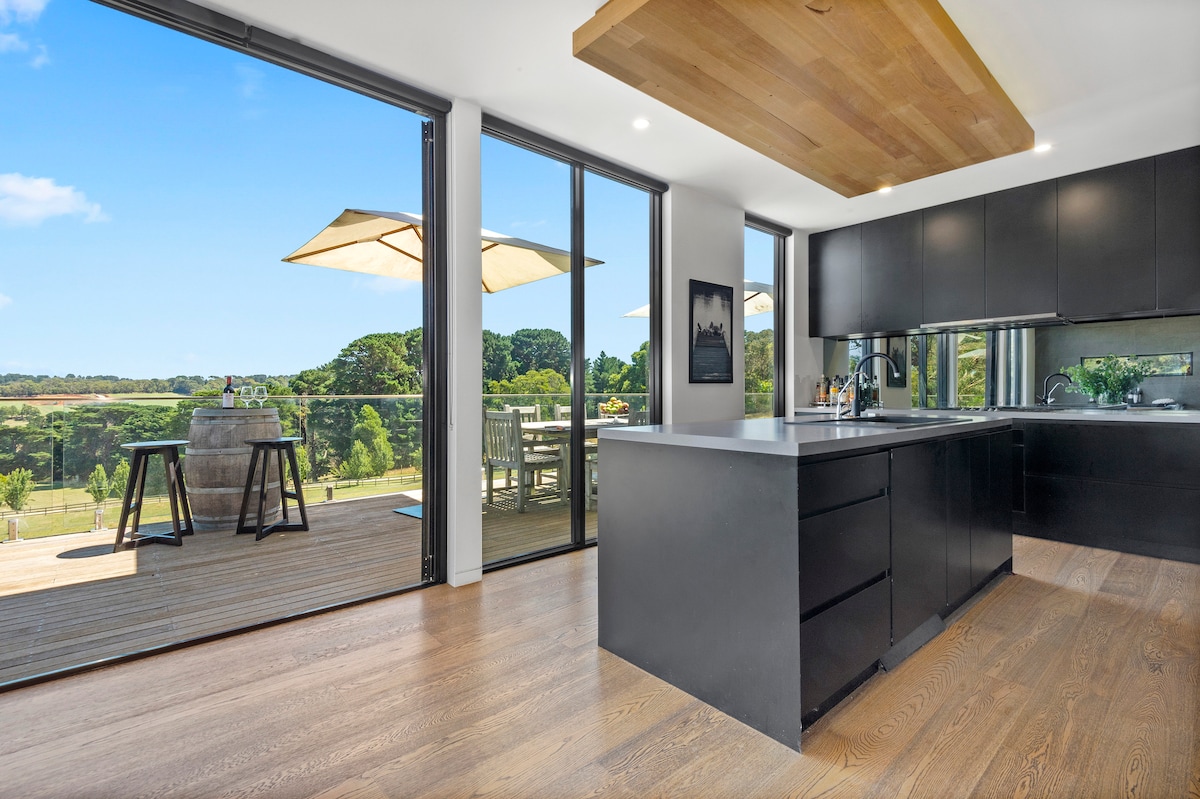 A modern kitchen features sleek black cabinetry and a spacious island, complemented by natural light from large sliding glass doors. An outdoor dining area is visible, showcasing a table and chairs beneath a canopy umbrella, with serene views of the surrounding countryside.