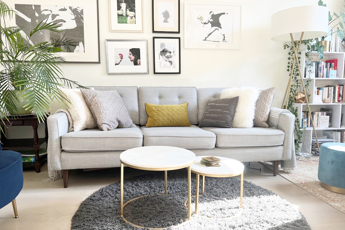 A light gray sofa is complemented by various textured throw pillows, positioned against a wall adorned with framed artworks. A round white coffee table is situated on a plush gray rug, with a small stack of magazines resting atop it. A green plant adds a natural touch.