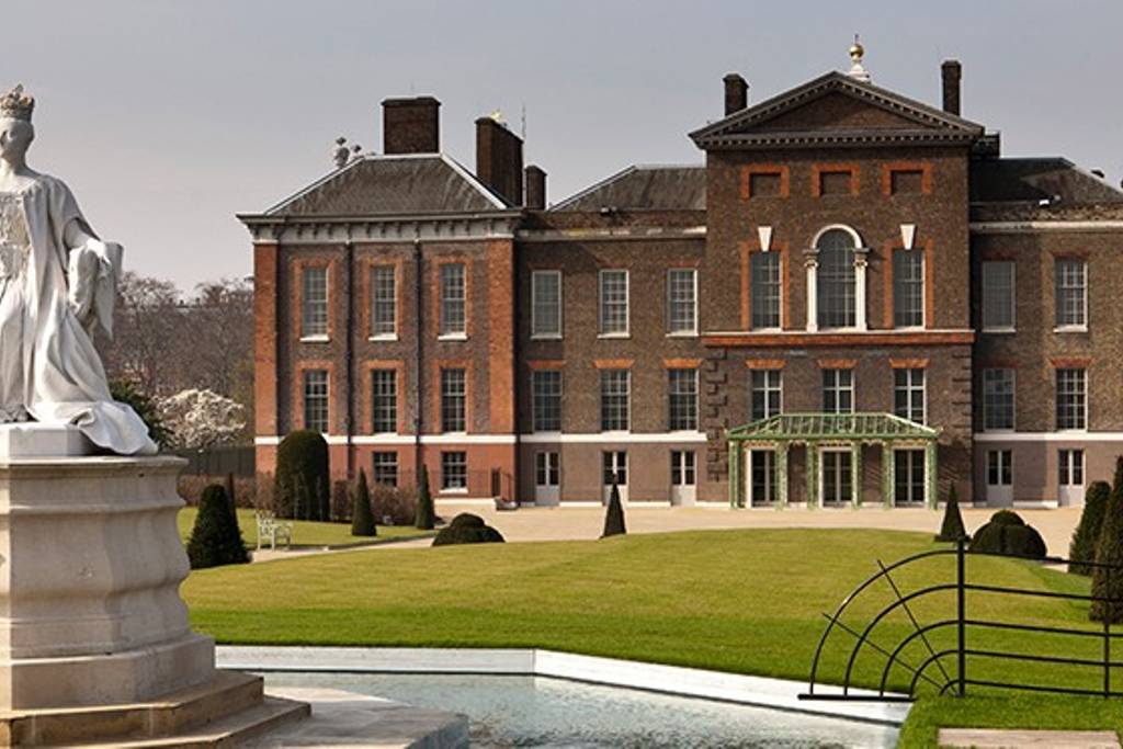 A grand historical mansion is displayed, featuring a symmetrical façade with large windows and a prominent entrance. Well-manicured lawns, shaped hedges, and a reflective pond complement the architectural details, while a statue stands in the foreground framing the property.