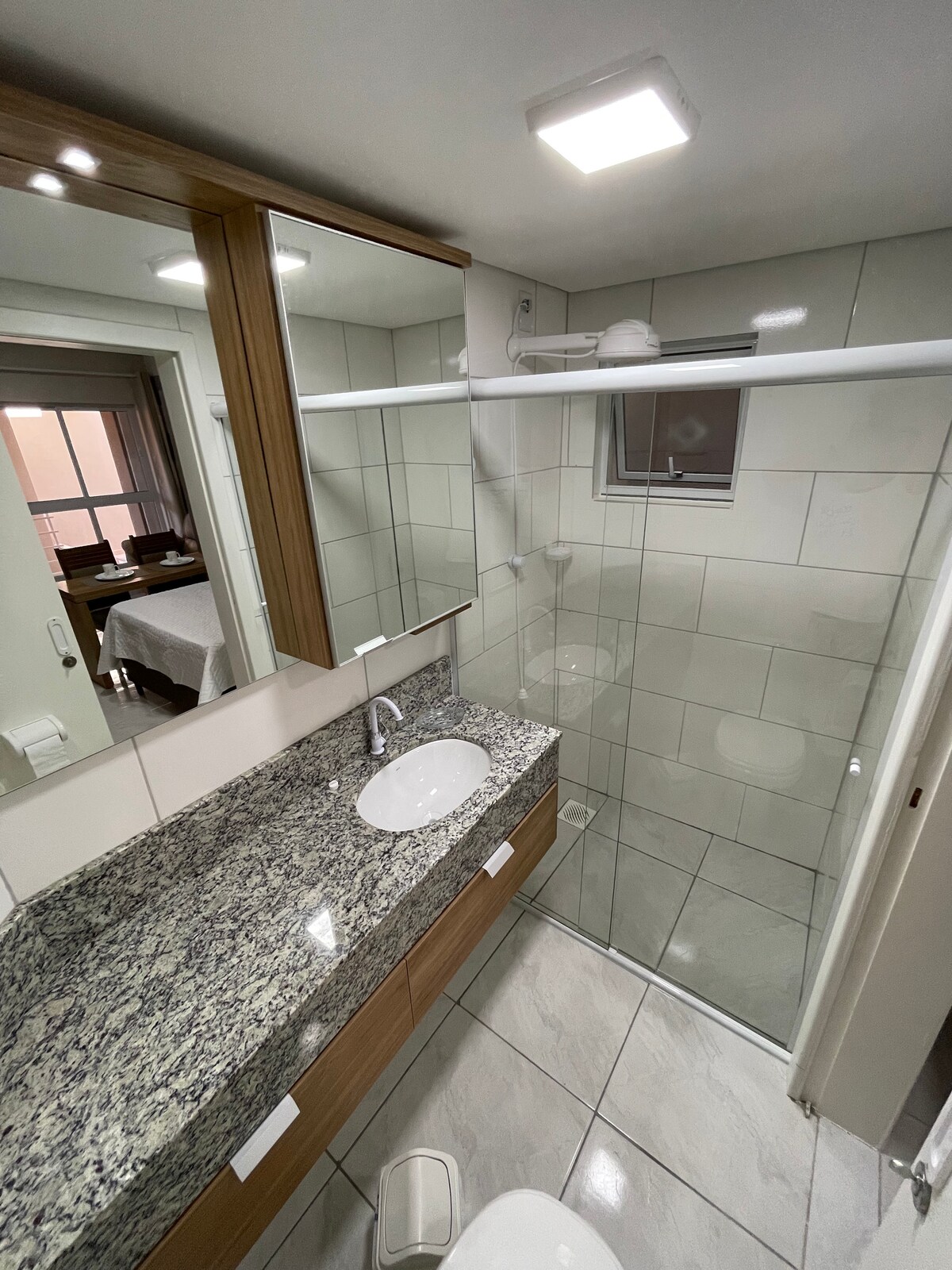 A modern bathroom features a spacious granite countertop with a round sink. Above, two mirrors reflect light, increasing brightness. A glass shower enclosure is visible, showcasing tiled walls and a clean design. Natural light enters from a nearby window, enhancing the inviting atmosphere.