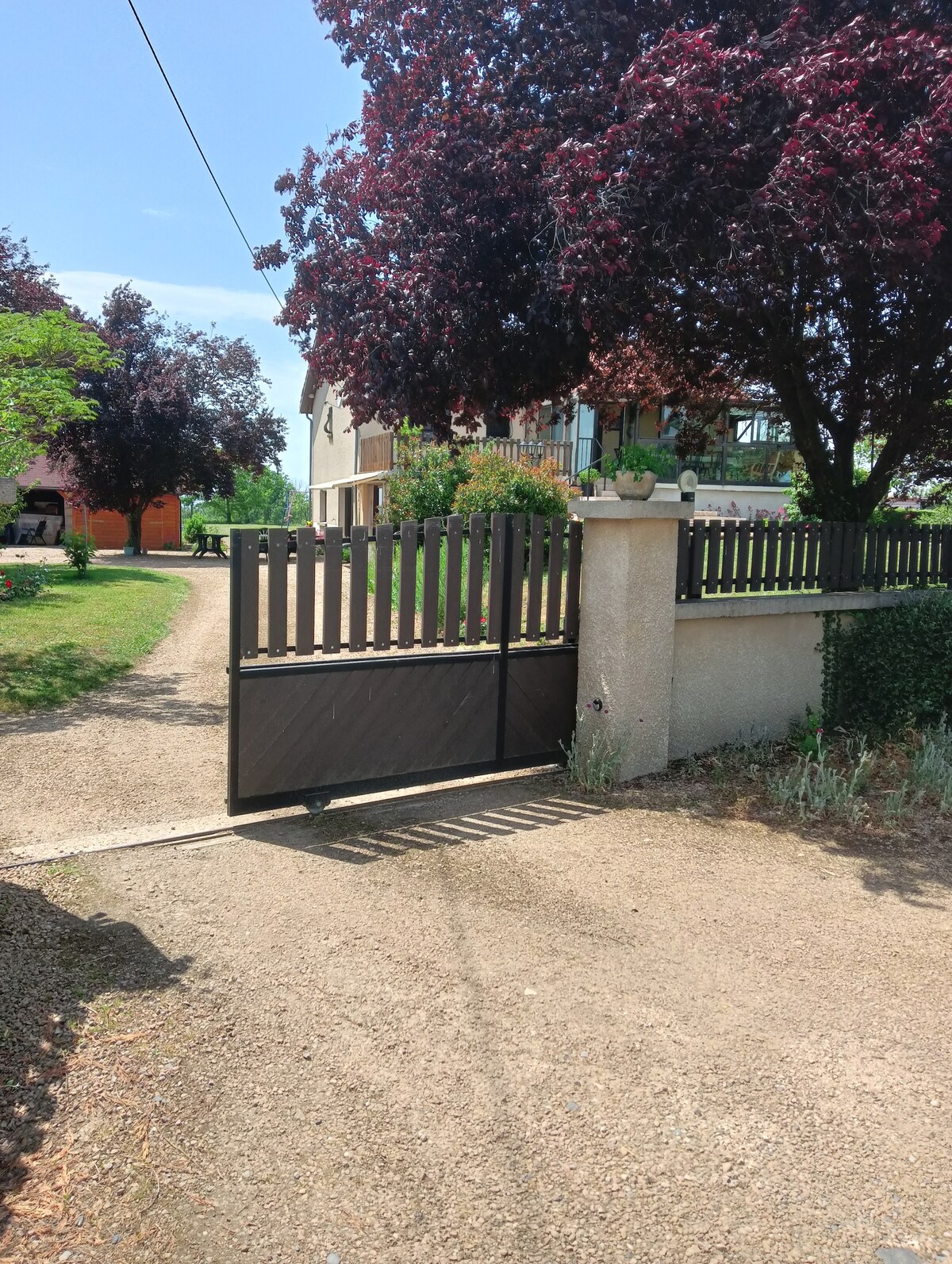 An automatic gate opens to a gravel driveway, surrounded by green trees and shrubs. A spacious property is visible in the background, featuring a light-colored building with large windows. The scene captures a welcoming entryway to the home.