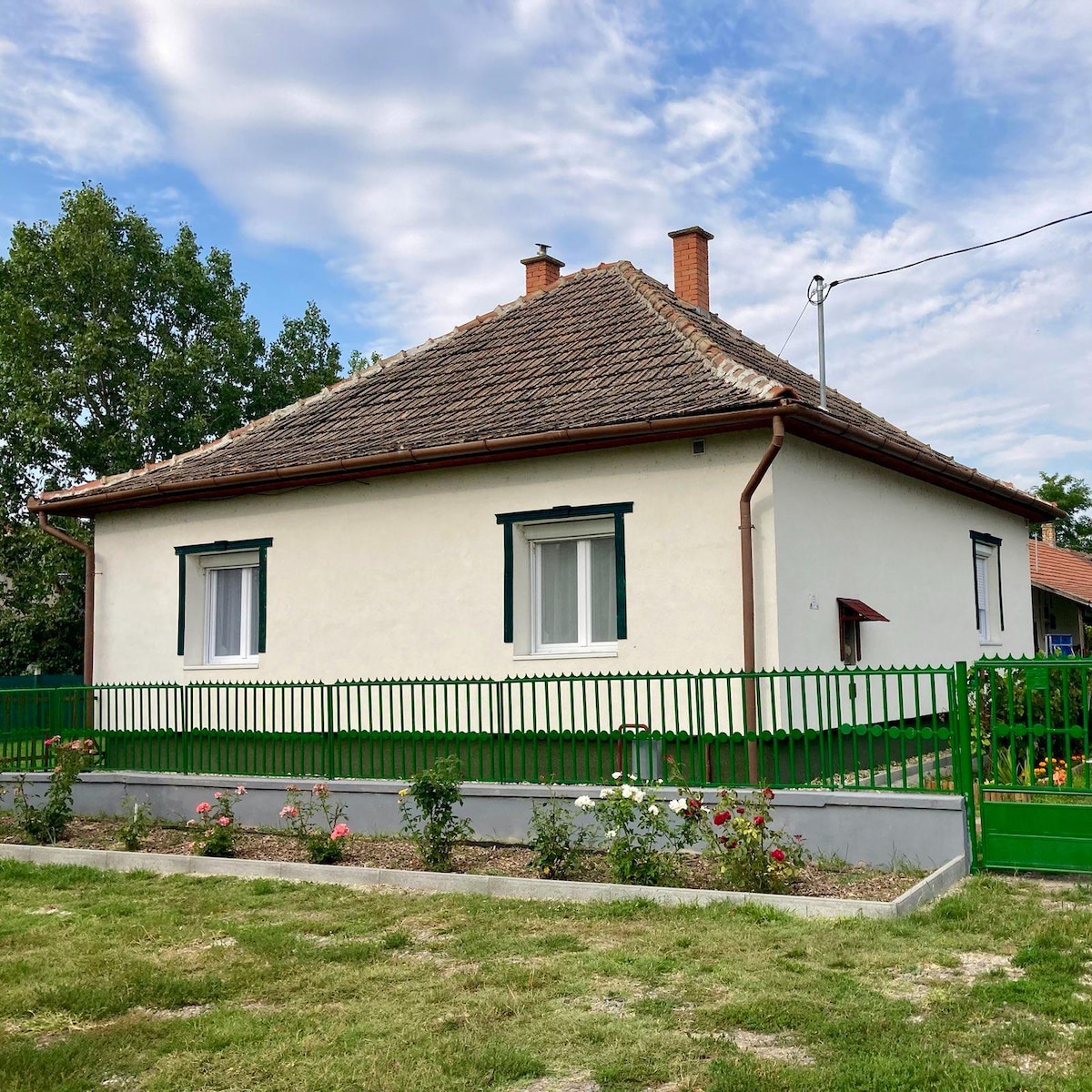 A single-story house is seen with a light-colored exterior and a traditional sloped roof. Green shutters accentuate the windows. A well-maintained garden with rose bushes is visible in front, surrounded by a low concrete wall and a green fence. The sky above is partly cloudy.