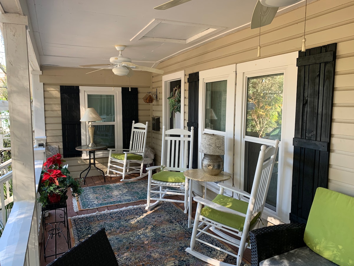 A welcoming front porch features several rocking chairs, a small table, and decorative plants. Light-colored shutters frame the windows, and a rug adds a cozy touch. Fans are visible on the ceiling, complementing the structure's inviting atmosphere.