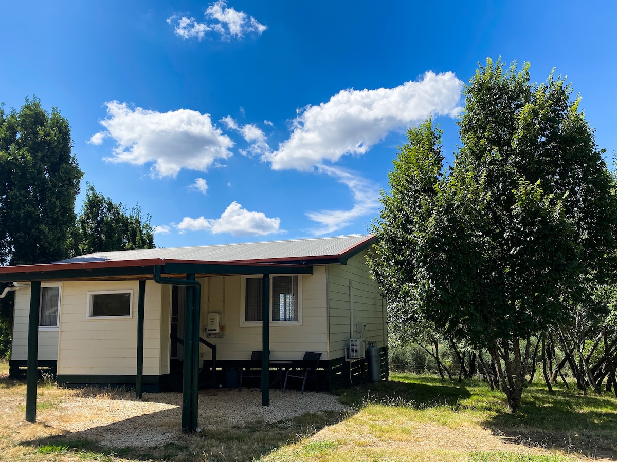 The cottage is set amidst greenery, featuring a light-colored exterior and a pitched roof. Large windows allow natural light to enter the interior. Nearby, two trees provide shade, and a gravel path leads to the entrance, illustrating a serene countryside setting.