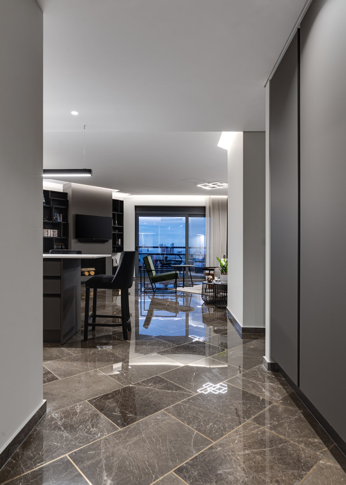 A view of the apartment's hallway leads towards an open living area, featuring sleek dark marble flooring. Light is reflected from the polished surfaces, while large glass doors provide natural light and access to a balcony with panoramic sea views.