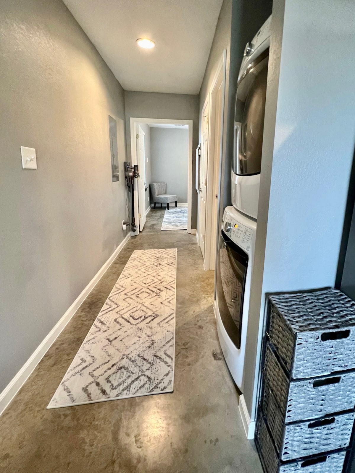 A narrow hallway is depicted, featuring a light-colored rug with a geometric pattern running along the floor. On one side, a stacked washer and dryer unit is positioned, while the opposite side showcases neatly organized storage bins. Soft lighting illuminates the space.
