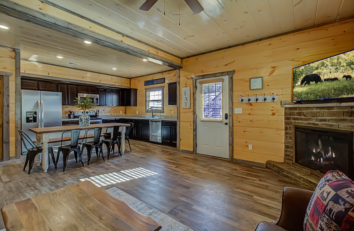 An open concept living area features wood-paneled walls and a wooden ceiling. A modern kitchen with stainless steel appliances overlooks a dining table surrounded by black metal chairs. A fireplace is positioned next to the entrance door, while a flat-screen TV is mounted on the wall.