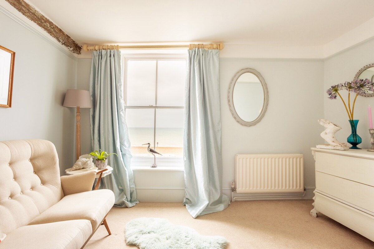 A light-filled room features a large window with sea views, dressed in soft blue drapes. A comfortable beige sofa is positioned opposite the window, complemented by a decorative mirror. A small decorative vase adds a touch of color to the neutral decor.
