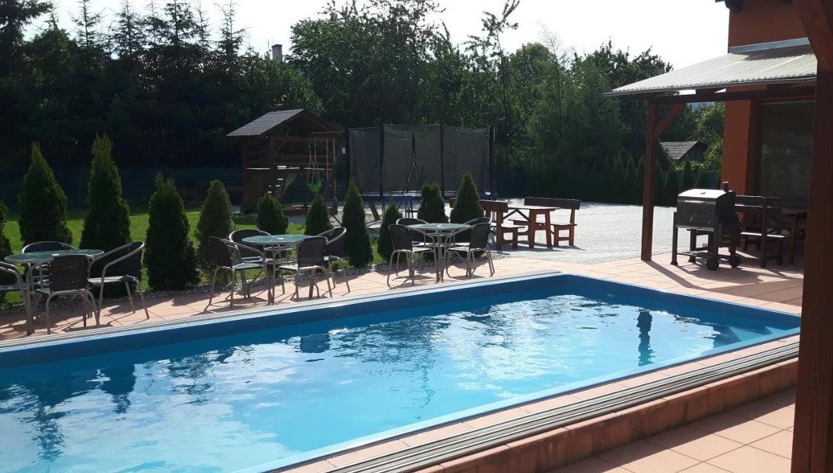 A clear swimming pool is surrounded by a tiled patio, featuring several tables and chairs for outdoor seating. Lush greenery and small coniferous trees line the area, providing a natural backdrop. A shaded structure is visible to the side, adding to the outdoor amenities.