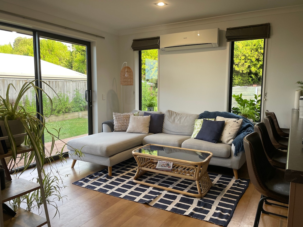 A comfortable living area features a light-colored sectional sofa with decorative pillows. A rattan coffee table sits on a geometric rug. Large glass doors provide views of the garden, while natural light fills the space through windows on either side.