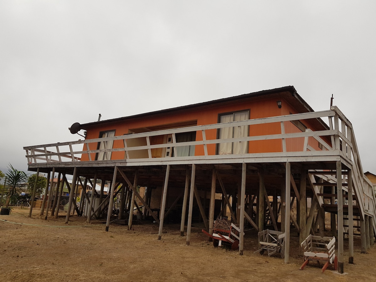 A raised house with a vibrant orange exterior is supported by sturdy wooden stilts, providing a unique view. Large windows are visible, allowing natural light to enter the living spaces. The surrounding area features open grounds, with some outdoor seating arrangements in view.