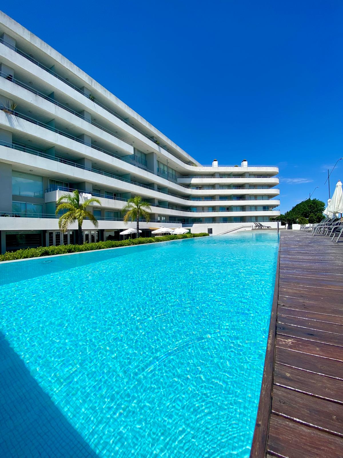 The outdoor swimming pool is framed by a wooden deck and lush greenery. Modern white apartment buildings rise in the background under a clear blue sky, reflecting a tranquil atmosphere. Sun loungers are positioned along the deck for relaxation.