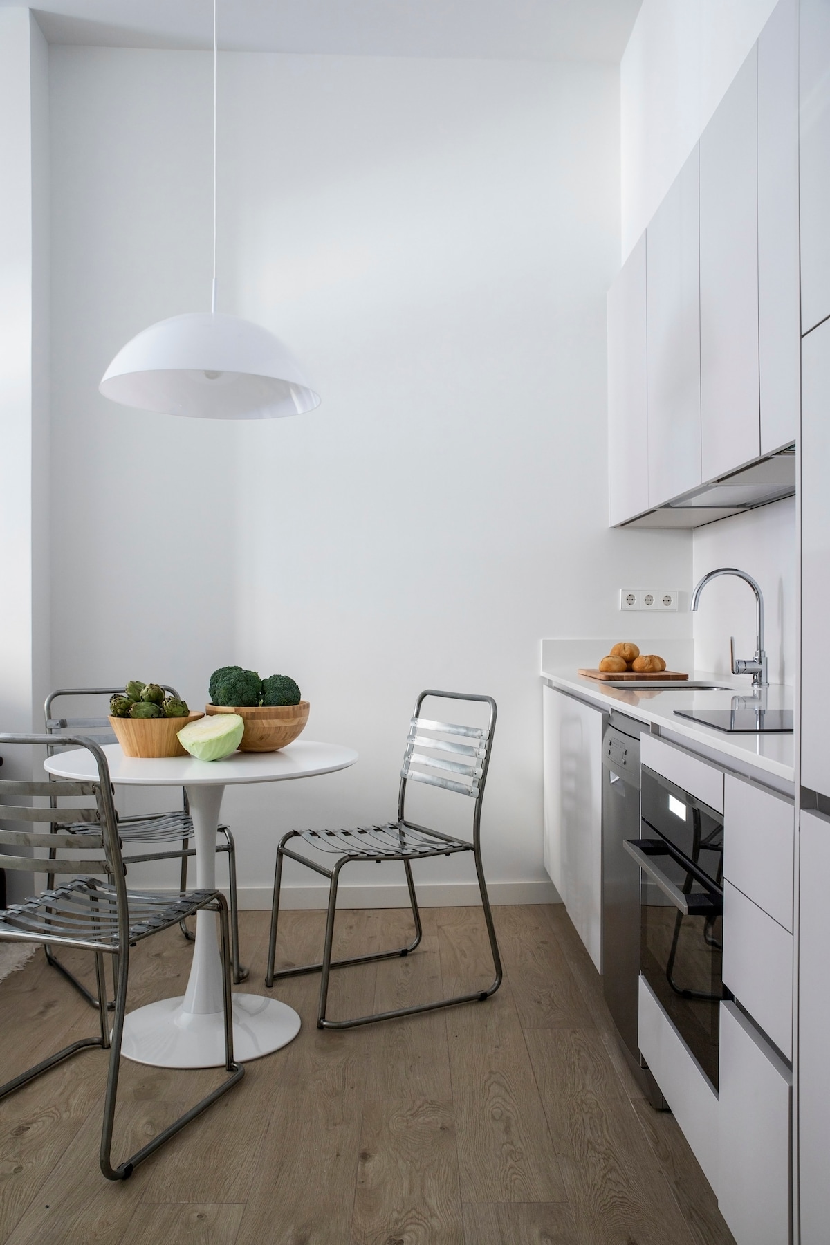 A bright kitchen space features a round white table paired with metallic chairs. Fresh vegetables are arranged in bowls atop the table, while modern kitchen appliances are integrated into the cabinetry. Light wood flooring complements the minimalist design.