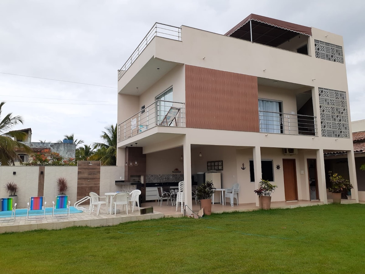 A modern two-story building is surrounded by green grass and features large windows. An outdoor seating area is visible, along with a well-maintained pool. A covered terrace on the upper floor offers additional leisure space, complemented by palm trees in the background.