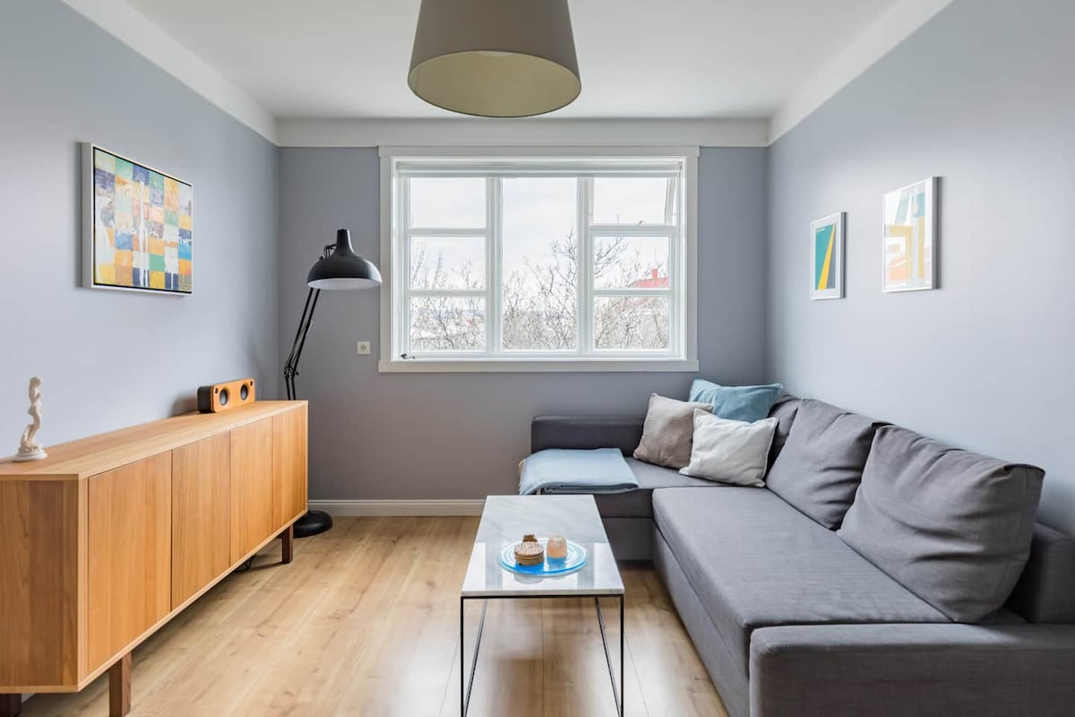 A light-filled living space features a gray sectional sofa accented with colorful pillows. A wooden sideboard adds warmth, while a contemporary coffee table sits in front. Pictures adorn the walls, and a floor lamp provides additional lighting near the window.