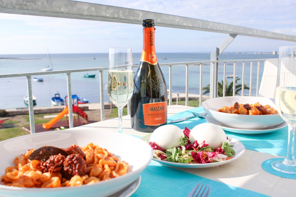 An inviting dining setup is presented on a balcony overlooking the sea. A bottle of Prosecco rests alongside elegant glasses, with two plates of pasta and a fresh salad arranged on a table adorned with a turquoise cloth. The tranquil waterfront features boats and a playground in the background.