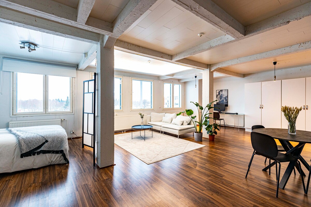 A spacious living area features large windows allowing natural light to fill the space. White walls and wooden flooring complement the modern furniture, including a comfortable sofa, dining table, and occasional plants. A cozy sleeping area with a large bed is visible in the corner.