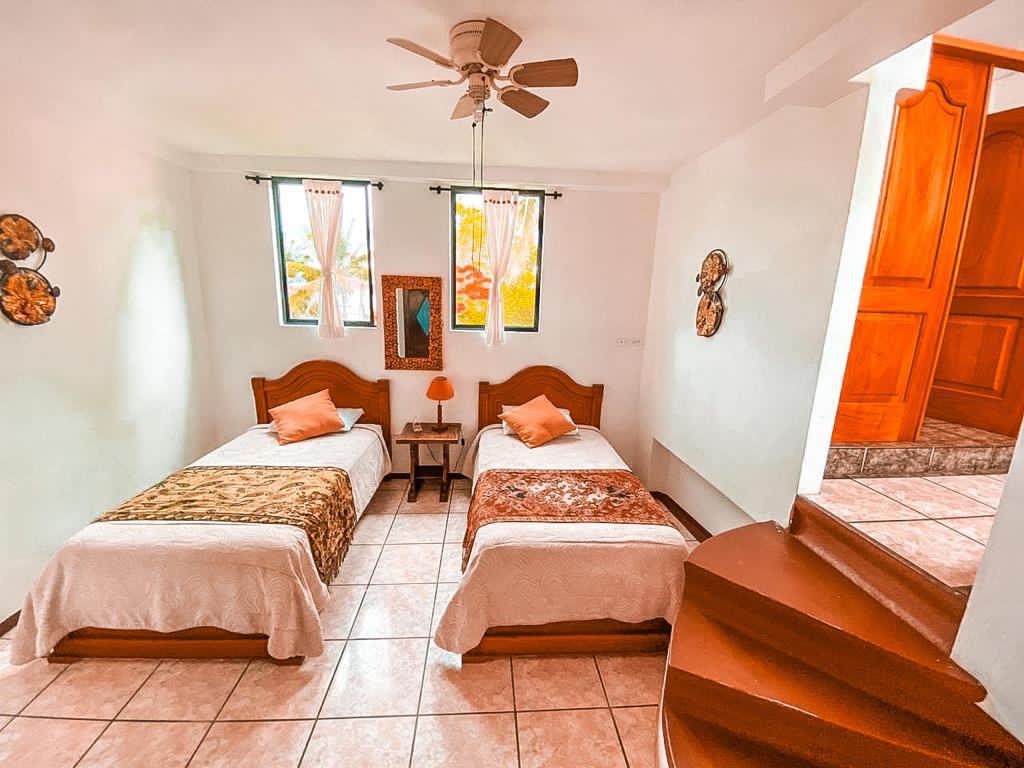 A bright room featuring two twin beds dressed in neutral bedspreads, complemented by decorative pillows. Natural light enters through three windows, illuminating the space. A small nightstand with a lamp is positioned between the beds, and a staircase leads to an upper level.