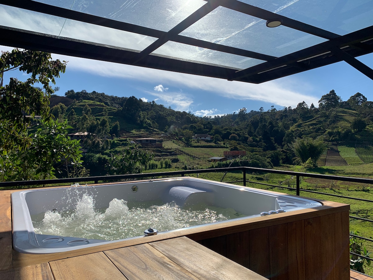 An outdoor jacuzzi, filled with bubbling water, is positioned under a glass roof. A panoramic view of lush green hills and fields extends in the background, providing a peaceful natural setting. The wooden deck offers a smooth contrast to the vibrant landscape.