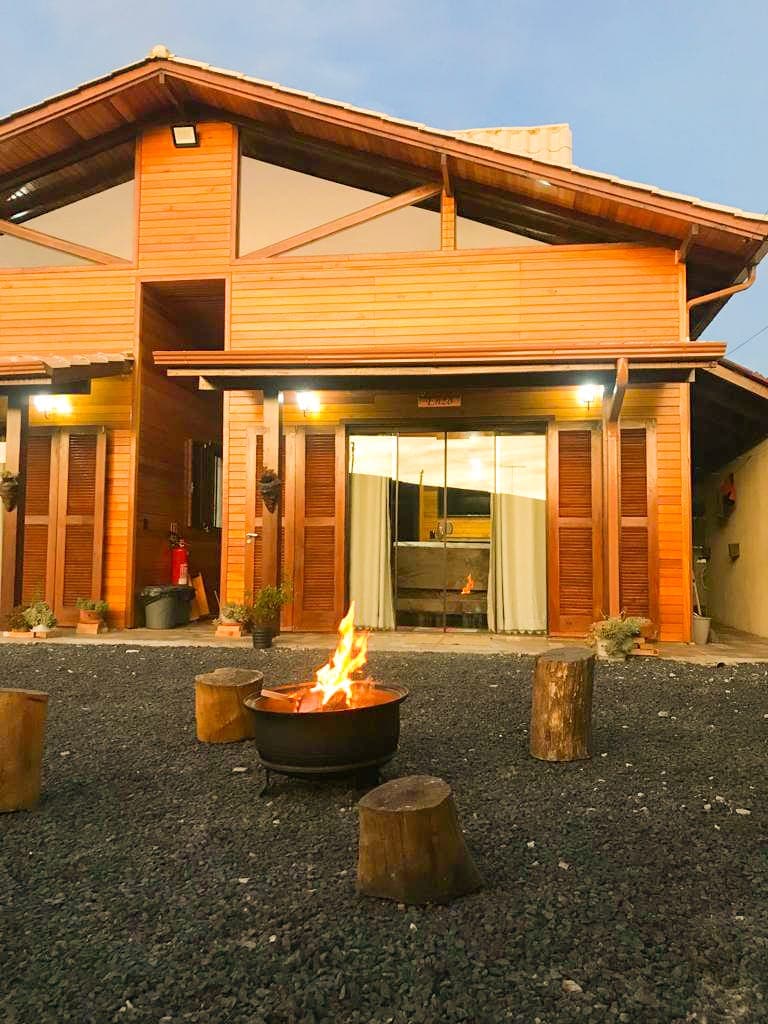 An exterior view of the wooden chalet features a cozy fire pit surrounded by log seating. Large glass doors provide a glimpse into the inviting interior, which is well-lit and welcoming. The rustic architecture is complemented by gravel landscaping.