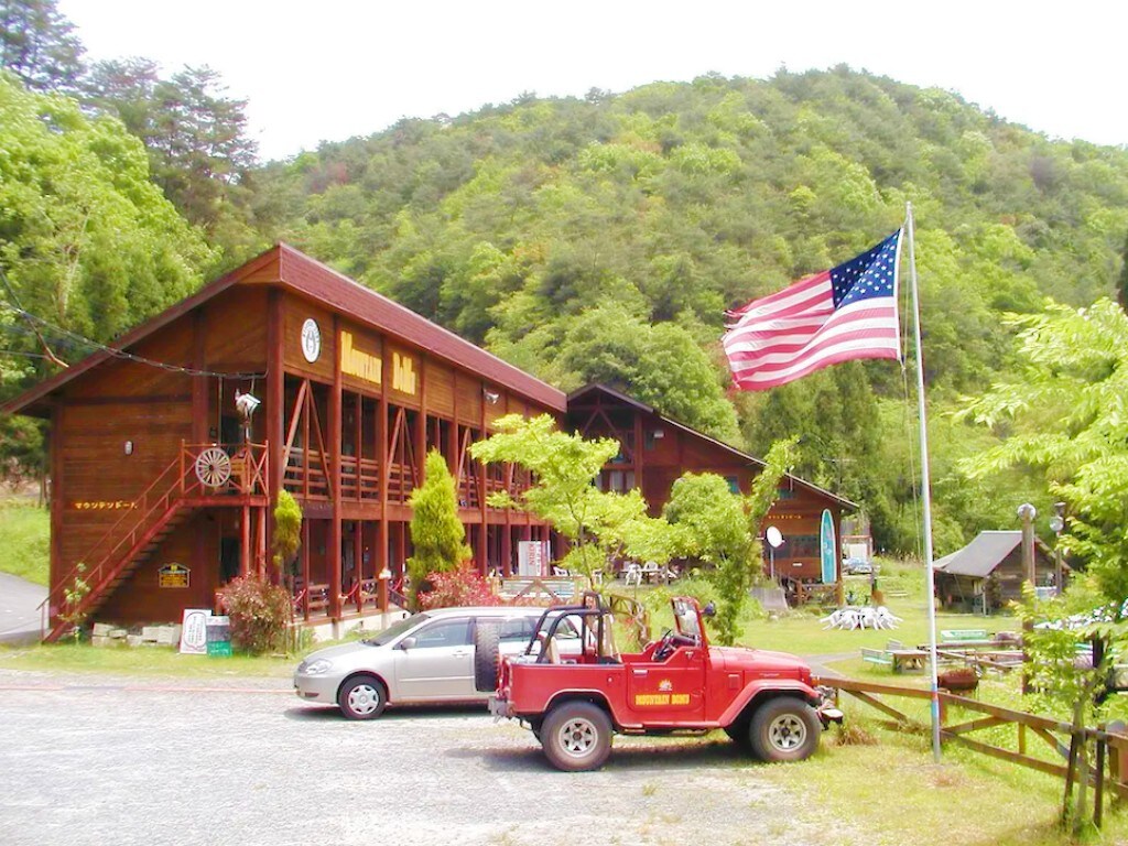 A wooden two-story structure is surrounded by lush greenery, with an American flag prominently displayed in front. Vehicles, including a red off-road car and a silver sedan, are parked in the gravel lot. The natural landscape provides a peaceful backdrop.
