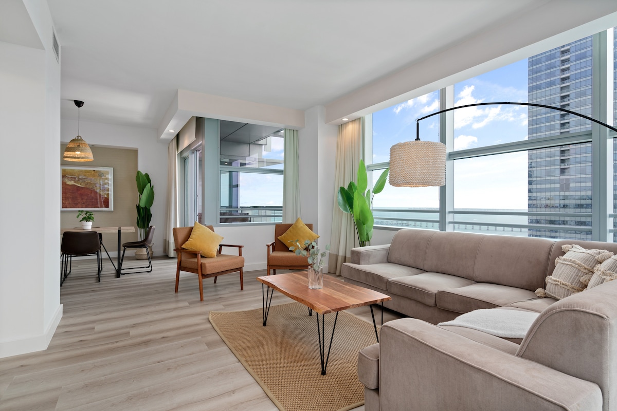The living area features large windows offering expansive views of the city and Biscayne Bay. A modern sofa is positioned across a wooden coffee table, surrounded by two stylish chairs. A light rug adds warmth to the space, complemented by soft lighting overhead.