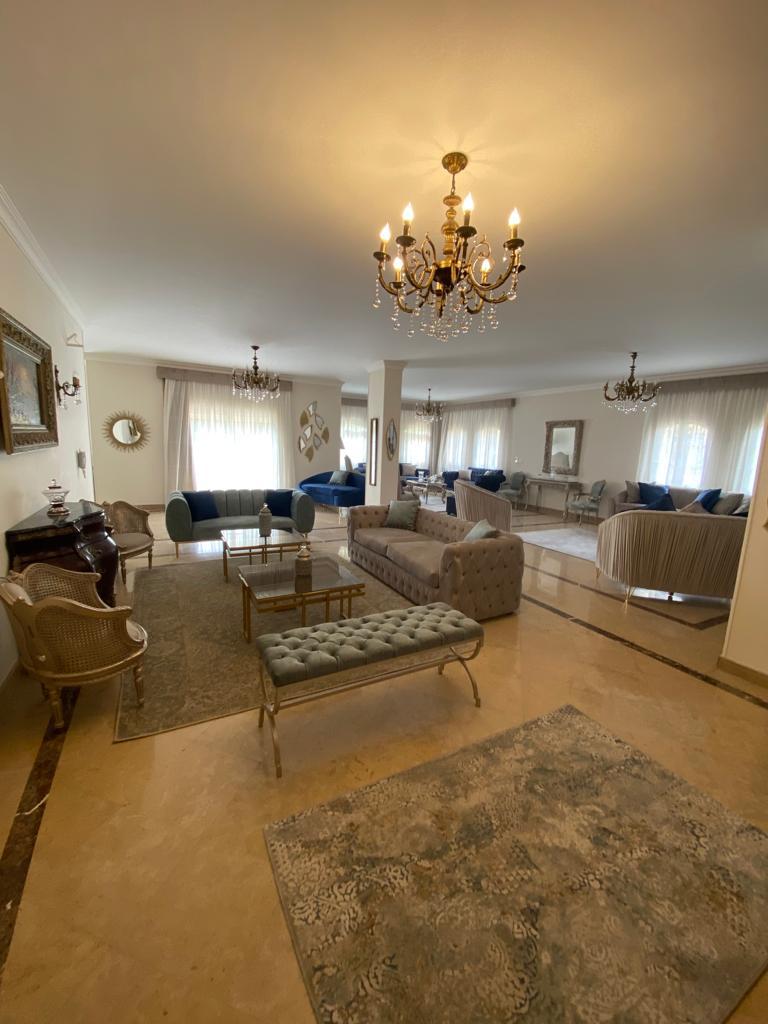 A spacious living area features elegant furnishings, including a tufted bench and a glass coffee table. Soft, neutral tones are complemented by plush blue sofas. Natural light filters through sheer curtains, highlighting decorative art and an ornate chandelier that adds sophistication to the space.